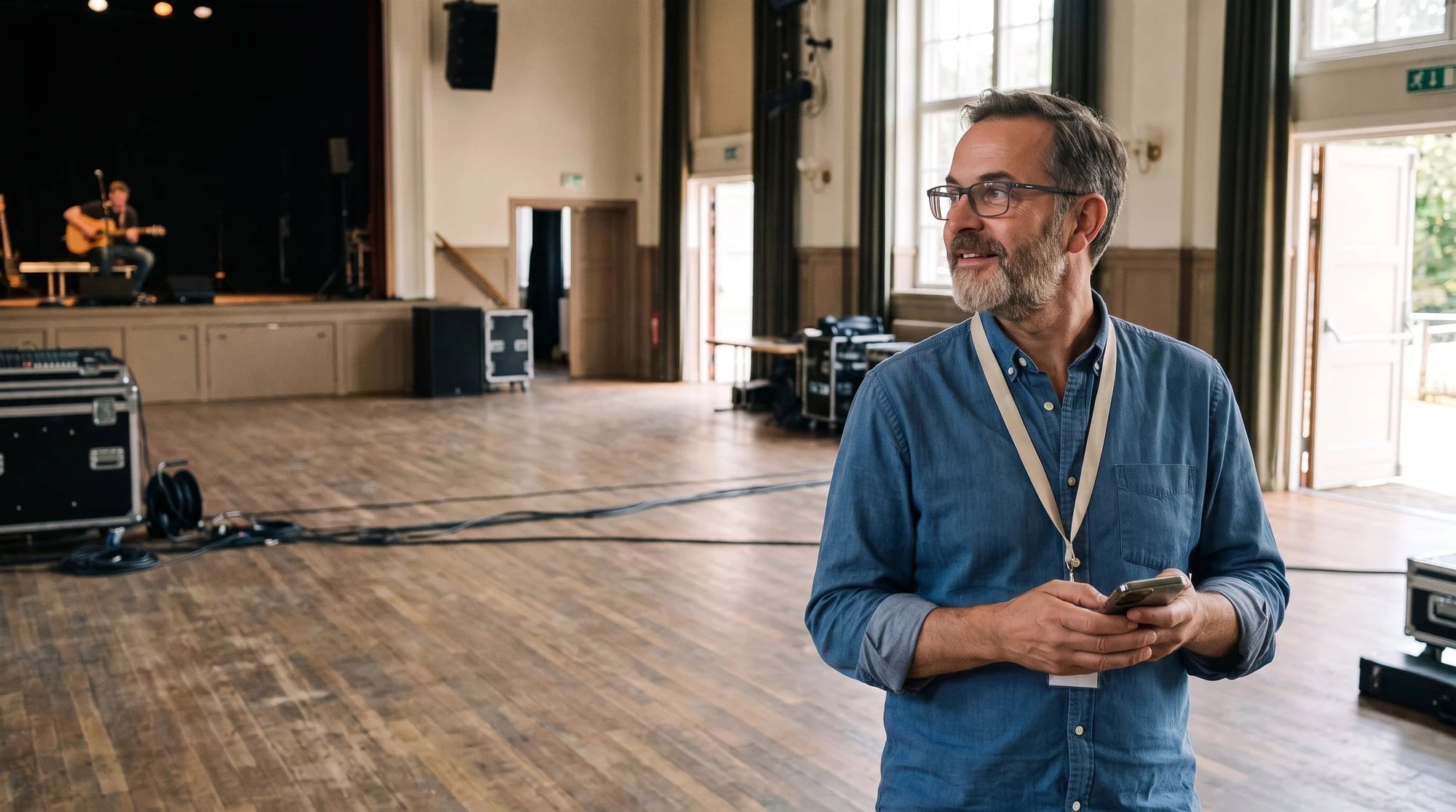 Konzertveranstalter mit Brille und Lanyard steht beim Soundcheck in der leeren Halle, Handy in der Hand, und blickt nach links zur Bühne, wo ein Gitarrist spielt